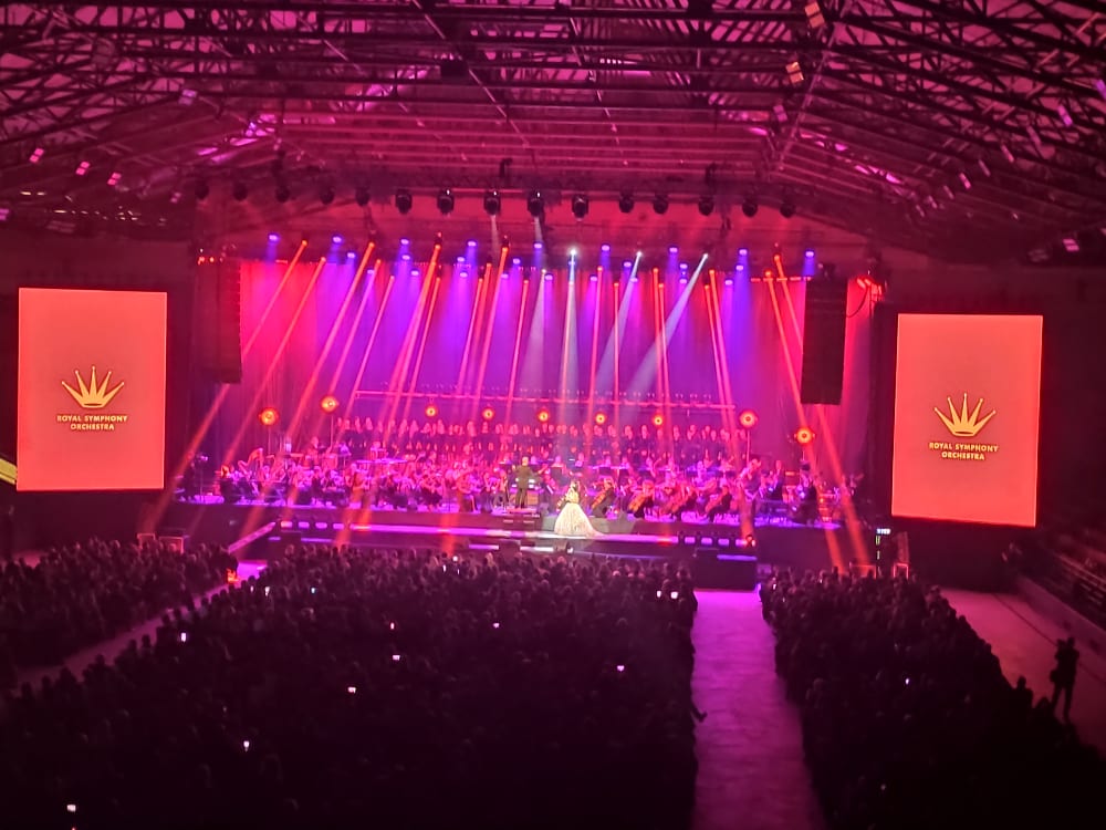 Koncert orkiestry symfonicznej i chóru na dużej scenie, oświetlony różowym i fioletowym światłem, z dwoma dużymi ekranami po bokach i publicznością na widowni.