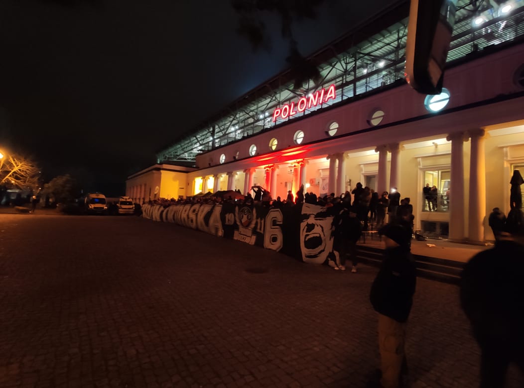 Nocny widok na fasadę stadionu Polonia Warszawa, z podświetlonym napisem 'POLONIA' i grupą kibiców z transparentami i flagami na placu przed wejściem.